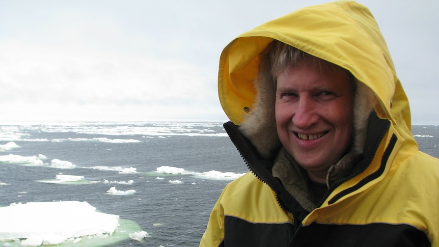 A man smiling in front an ocean with icebergs in a yellow jacket with the hood up