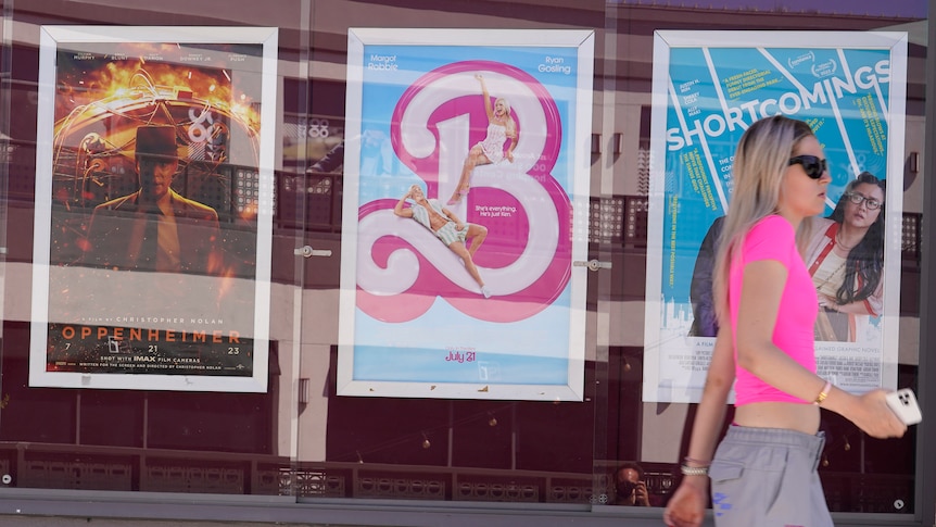 A blonde woman in a pink shirt walks past posters for Oppenheimer and the Barbie movie