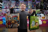 A small smiling boy holds up two show bags with colour and lights behind him.