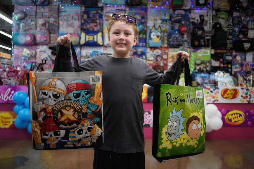 A small smiling boy holds up two show bags with colour and lights behind him.