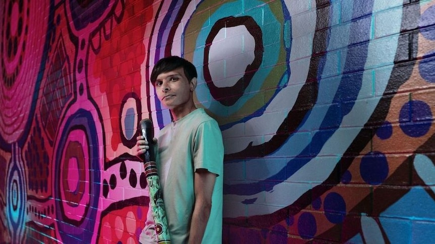 Yugambeh language expert Shaun Davies pictured against a graffitied wall, holding a didgeridoo
