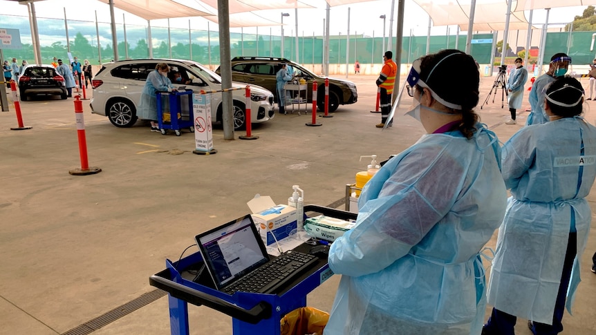 Healthcare workers in full PPE look on as other workers speak to people at car windows at a drive-through vaccination site.