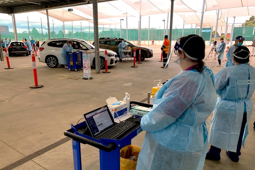 Healthcare workers in full PPE look on as other workers speak to people at car windows at a drive-through vaccination site.