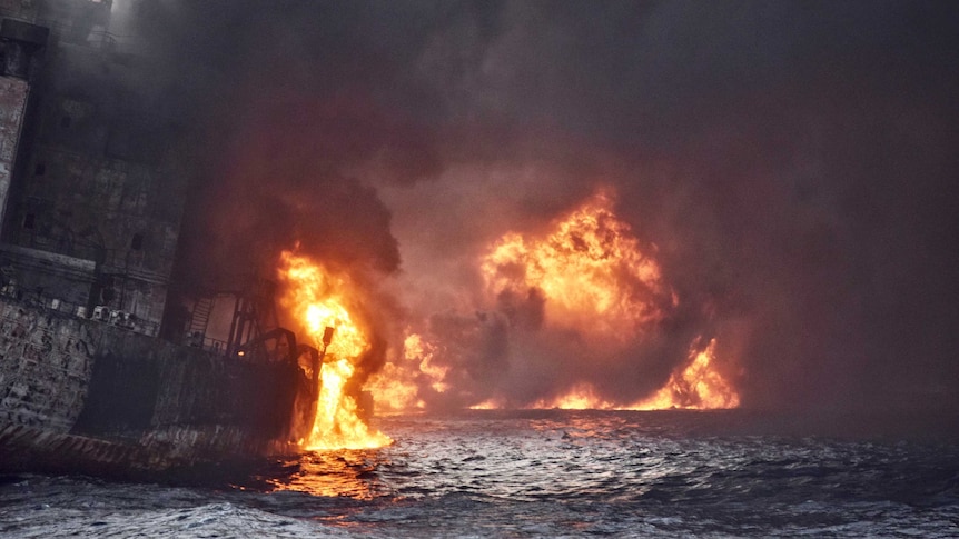 Heavy smoke pours from a ship ablaze in the ocean.