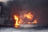 Heavy smoke pours from a ship ablaze in the ocean.