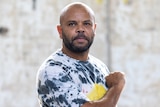 An Indigenous Australian man in an Aboriginal flag shirt holds a fist up to his heart, and looks at the camera.