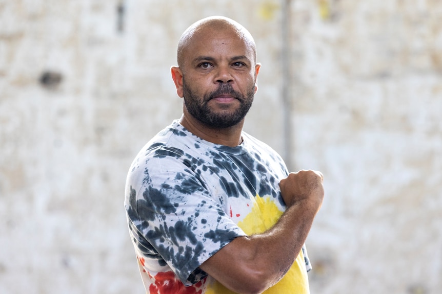 An Indigenous Australian man in an Aboriginal flag shirt holds a fist up to his heart, and looks at the camera.