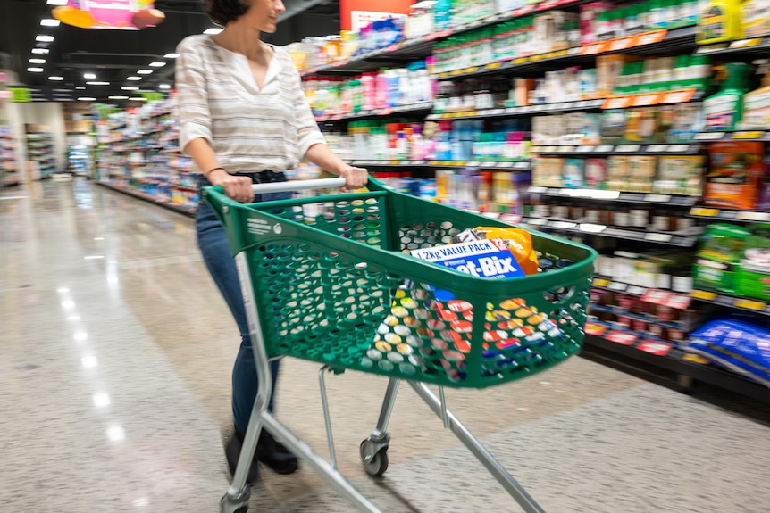 A woman wheeling a trolley down an aisle in a Woolworths store. The trolley has a few items in it.