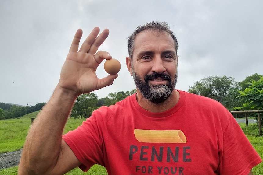 A man with a black and grey bear wearing a red shirt holds a round egg in his fingers.