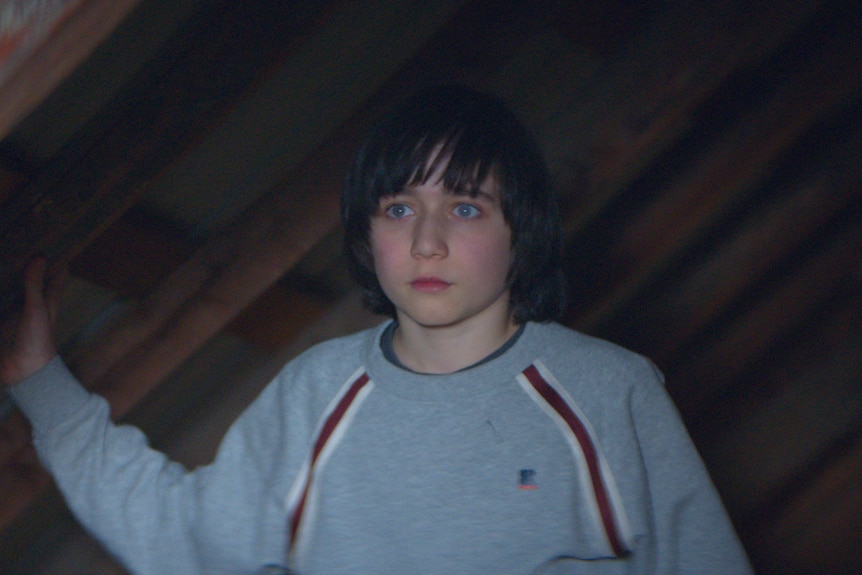 A film still of Milo Machado Graner, a young boy, standing in an attic, with a dazed expression.