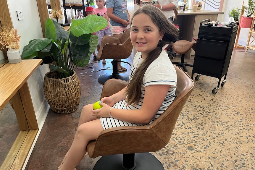 A young girl in a white dress with black stripes with long hair smiling and sitting in a chair.