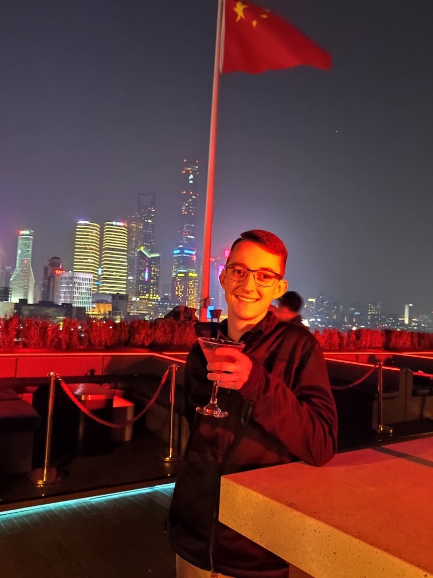 Christ Milne holds a martini glass at a rooftop bar in front of a Chinese flag, with a city skyline in the background.
