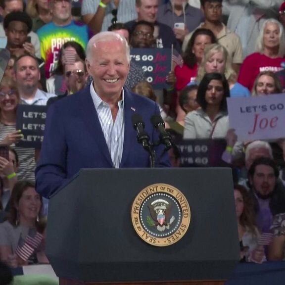 Joe Biden smiling at a microphone and lecturn with a crowd behind him.