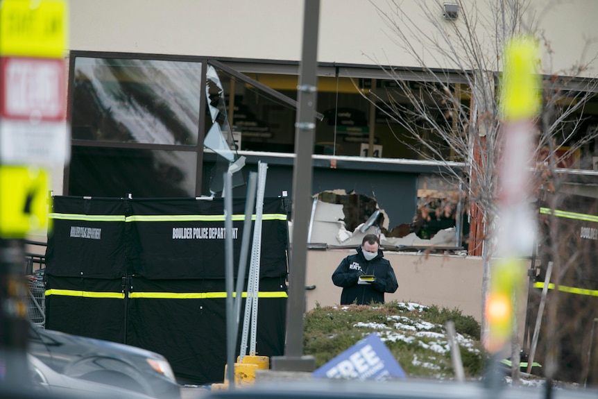 A police officer stands outside a building with shattered windows.