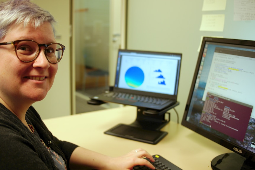 A woman wearing glasses sitting in front of a computer, smiling at the camera.