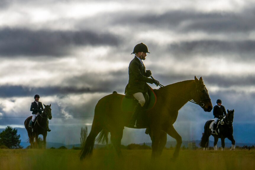 Horse riders are partially silhouetted by bands of sunlight glowing through clouds in a green field.