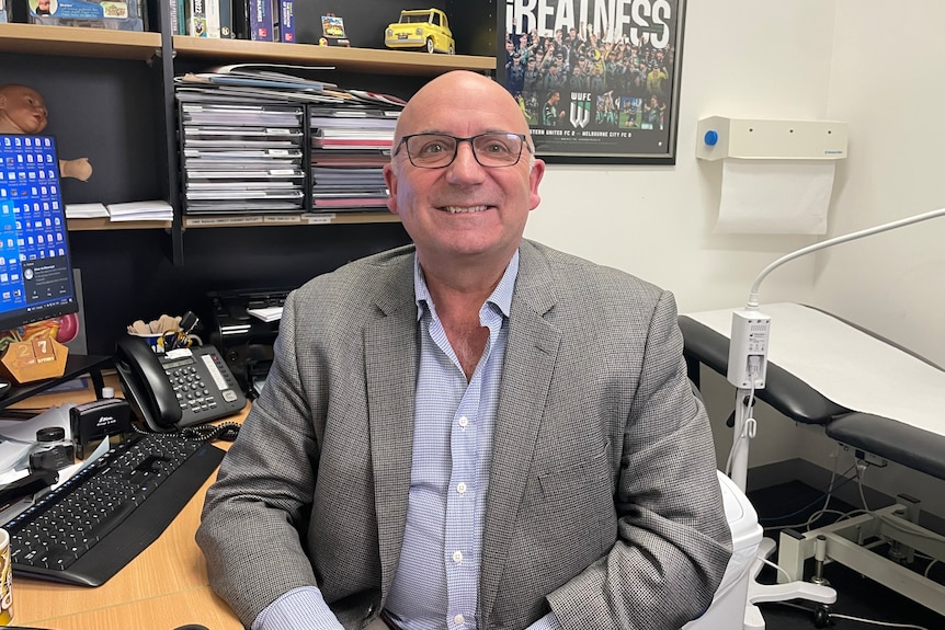 A man sitting at a desk, smiling and wearing glasses and a blazer.