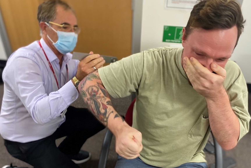 A researcher, wearing a mask measures a man's shoulder mobility, man covers mouth in shock.