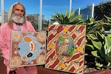 Stanley Geebung sits with two of his artworks outside in a garden.
