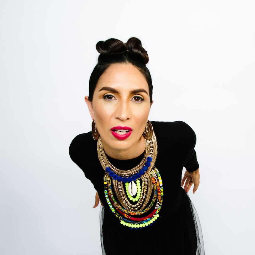 Maya Jupiter wearing a large beaded necklace standing in front of a white background leaning in towards the camera