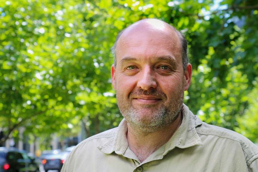 Craig Hinton stands beneath green tree canopy wearing khaki shirt and grey facial stubble.