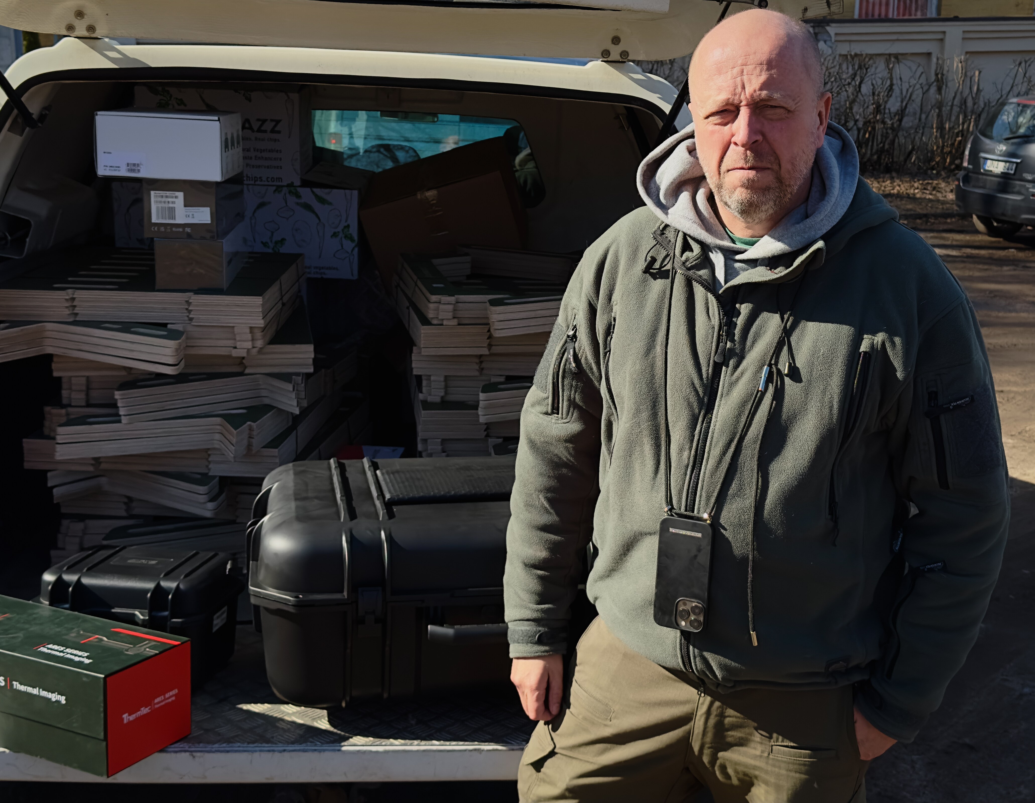 A man in a grey hoodie squints in the sunlight. He stands by an open car boot full of cases of drones