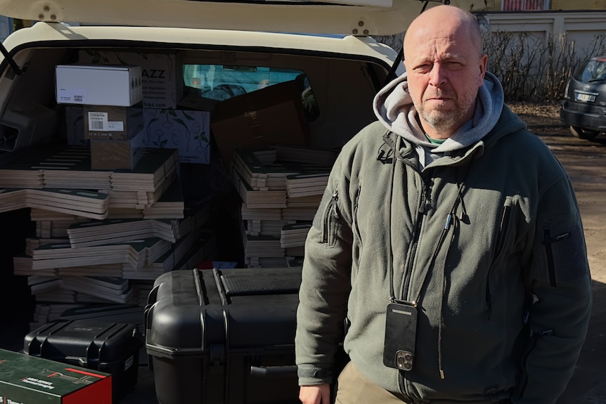 A man in a grey hoodie squints in the sunlight. He stands by an open car boot full of cases of drones
