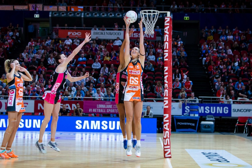 Netball players jump for the ball under the goal post.