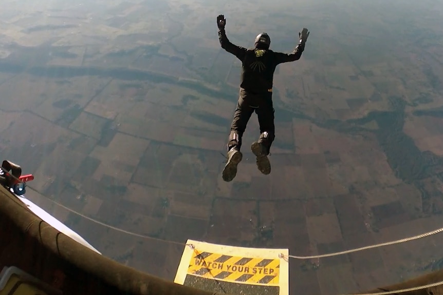 A view from above as a man jumps out of a vessel in the air with arms and legs spread, the ground in the distance below.