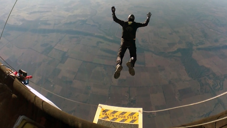 A view from above as a man jumps out of a vessel in the air with arms and legs spread, the ground in the distance below.