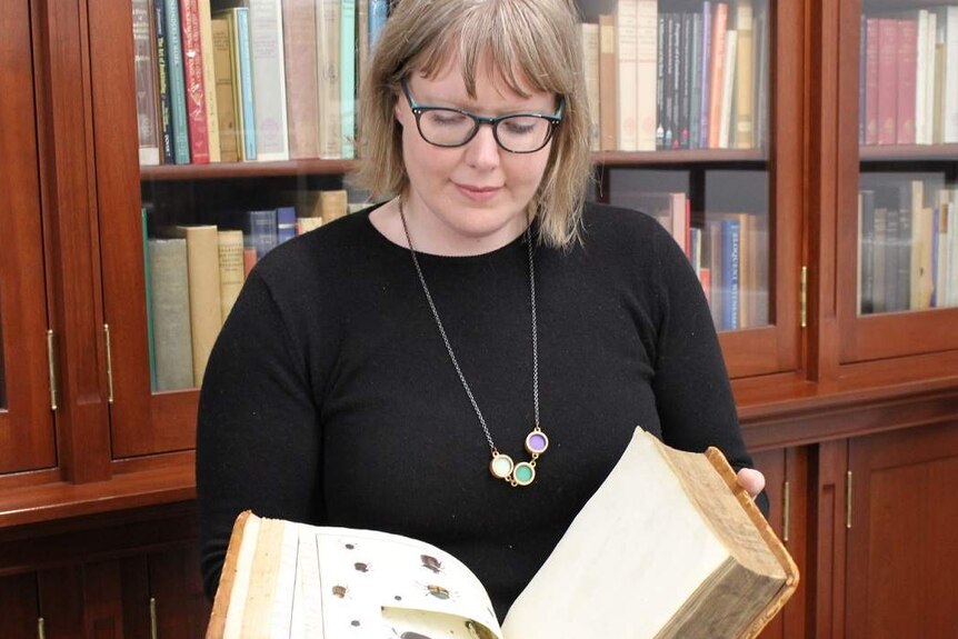 State Library historian Anna Welch holds open a 200-year-old book with it open to a page with a cut picture cut out.