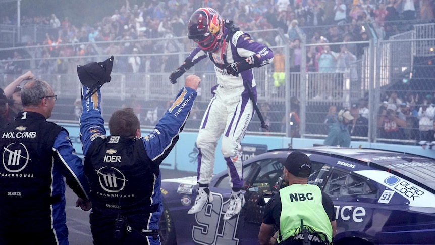 A NASCAR driver with his helmet and gear on jumps down from his car in pit lane after winning a big race.