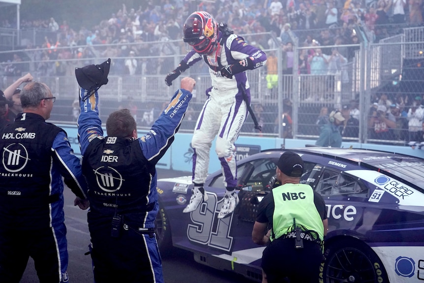 A NASCAR driver with his helmet and gear on jumps down from his car in pit lane after winning a big race.