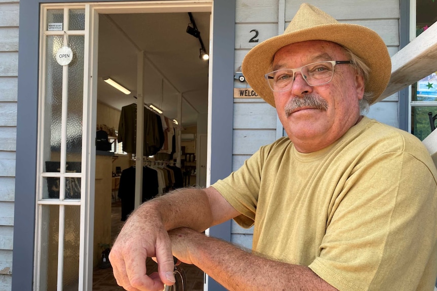 Brian Aitchison is sitting outside a cottage. He is wearing a hand and resting his arms on a walking stick