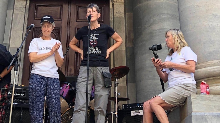 A woman speaking to a microphone with her hands on her hips between large grey columns