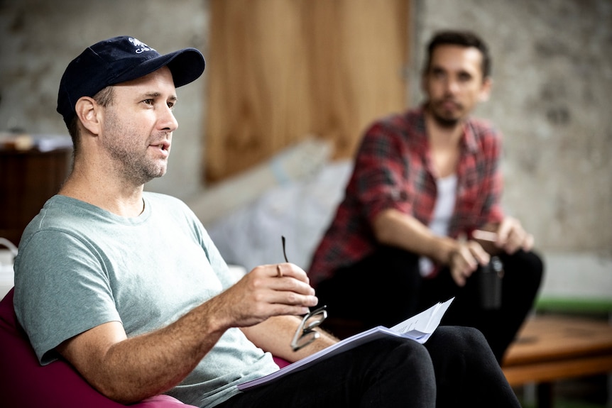 Eamon Flack sits on a couch on-set with a script and pen, talking to someone we can't see. In the background is an actor.