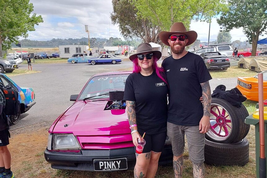 A man and woman in broad hats stand in front of a pink car. The woman has long pink hair.