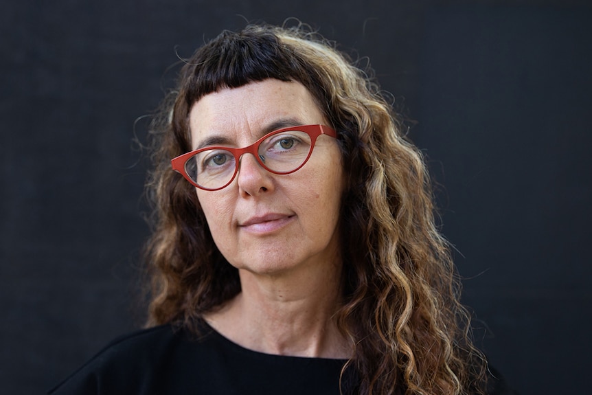 A woman wearing black t-shirt and red glasses looks towards camera and stands in front of a dark grey wall.