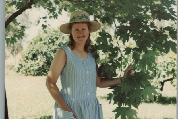 Penny Hill, wearing a hat and dress, stands under a tree.