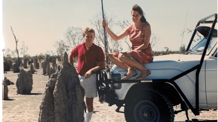 A woman sits on the bonnet of a large car while a man stands on the ground next to her in the outback