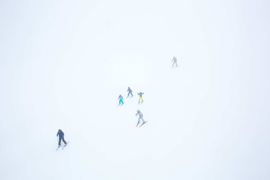 Skiers cut tiny figures against an expanse of white, as seen from above.