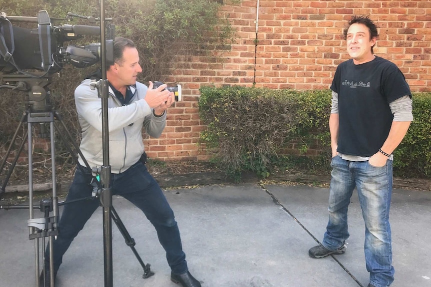 Greg Nelson standing next to video camera uses a stills camera to take a photo of Samuel Johnson.