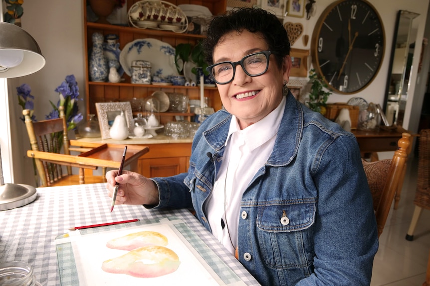 A woman with short dark hair sits at a table, smiling, holding a paintbrush.