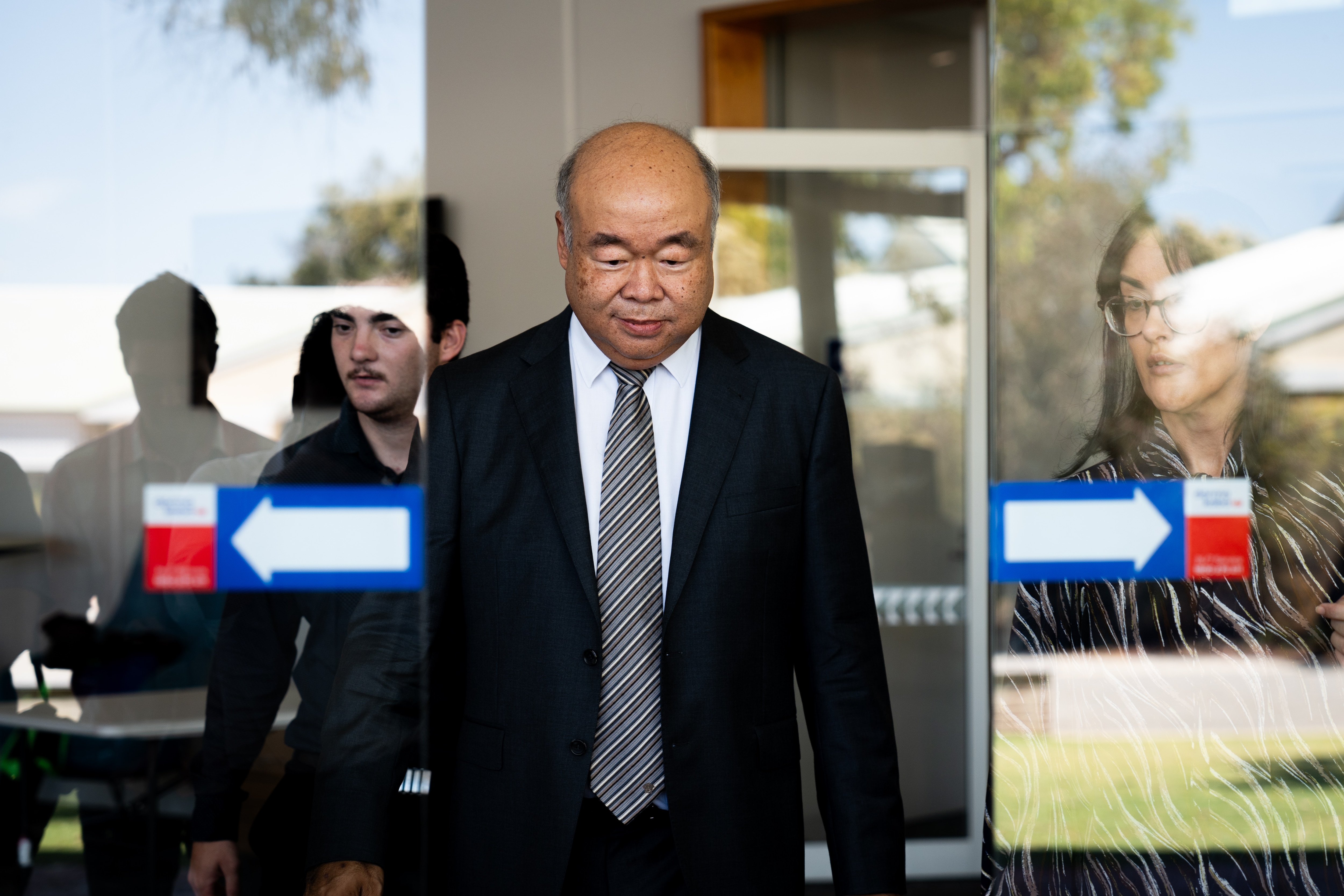A besuited man with eyes downcast exits through a sliding glass door