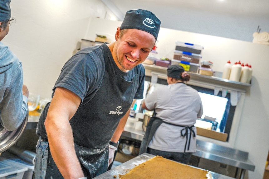 A man, wearing a baker's cap and apron, smiles as he works in a commercial kitchen