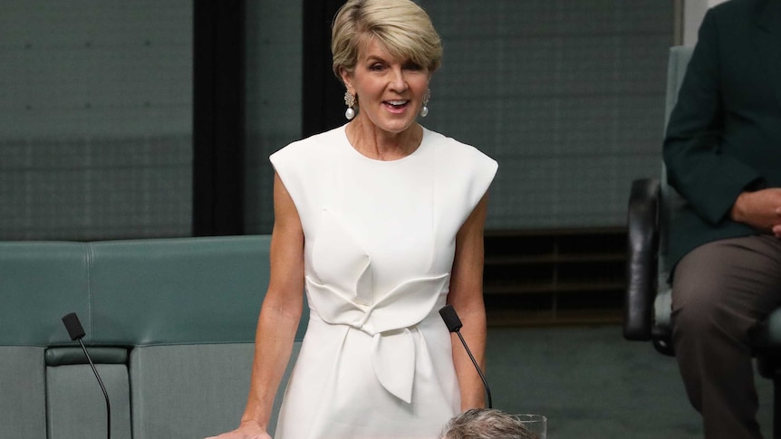Julie Bishop, standing and smiling, speaks to Parliament while wearing a white dress.