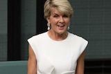 Julie Bishop, standing and smiling, speaks to Parliament while wearing a white dress.