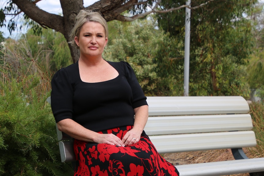 Tanya Steinbeck sits on a park bench with trees behind her wearing a black top and red skirt.