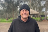 A man in a black jumper and black beanie looking at the camera, standing in a camping ground