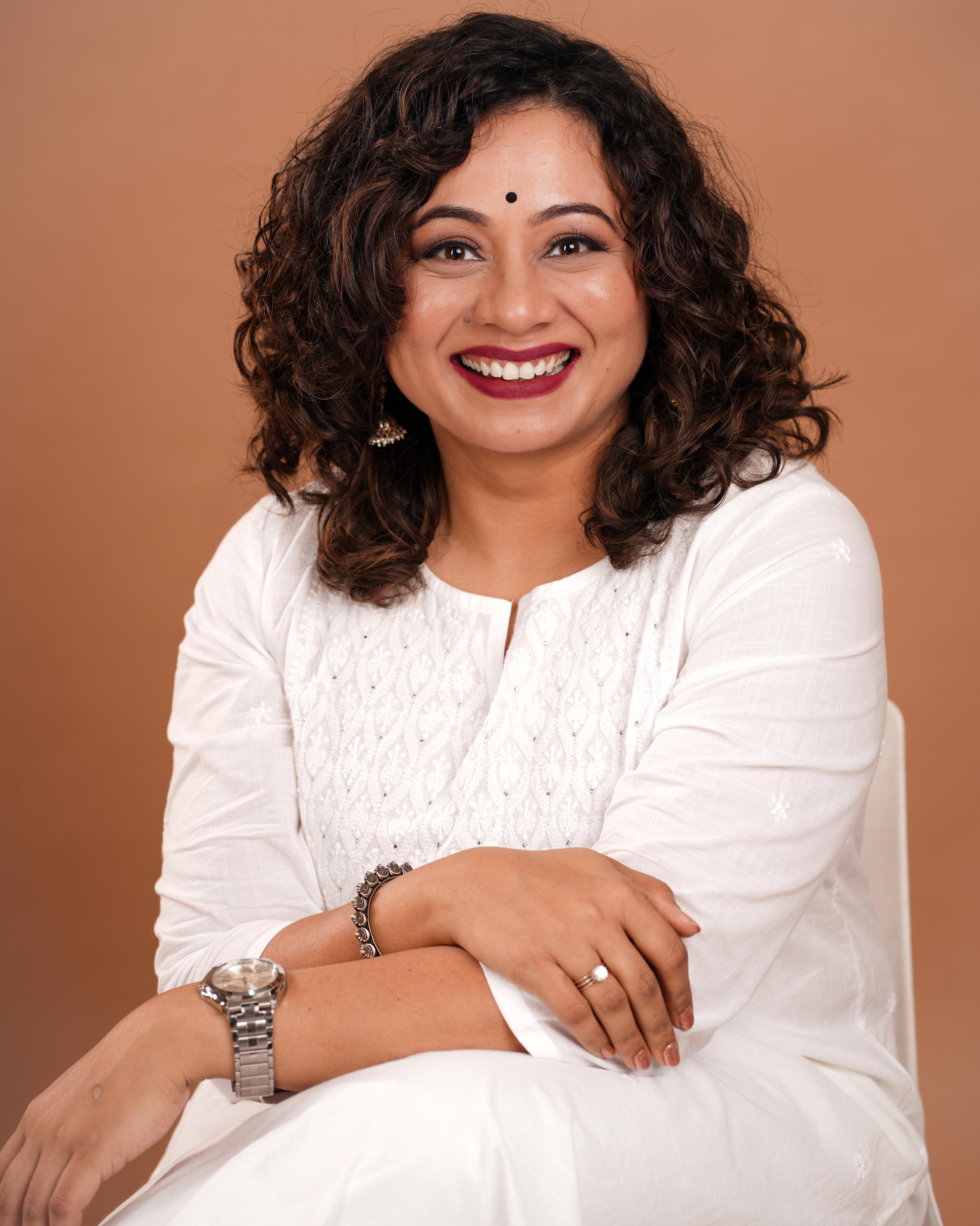 A woman in a white dress sits on a chair with her arms folded over.